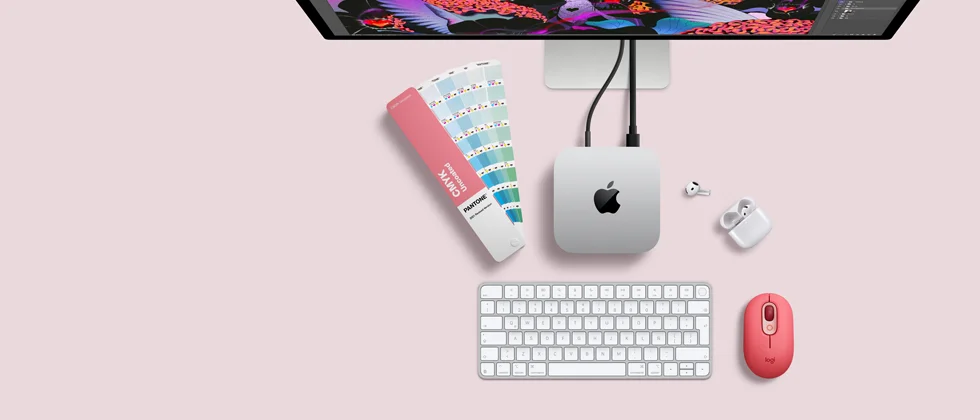 Mac mini, parte superior, aluminio color plata, esquinas redondeadas, logo de Apple en el centro en color negro brillante, conectada con un cable, monitor Studio Display parcialmente visible en la parte superior, AirPods 4, abiertos, muestra de una paleta de colores Pantone, teclado numérico y mouse
