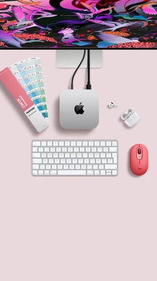 Mac mini, parte superior, aluminio color plata, esquinas redondeadas, logo de Apple en el centro en color negro brillante, conectada con un cable, monitor Studio Display parcialmente visible en la parte superior, AirPods 4, abiertos, muestra de una paleta de colores Pantone, teclado numérico y mouse